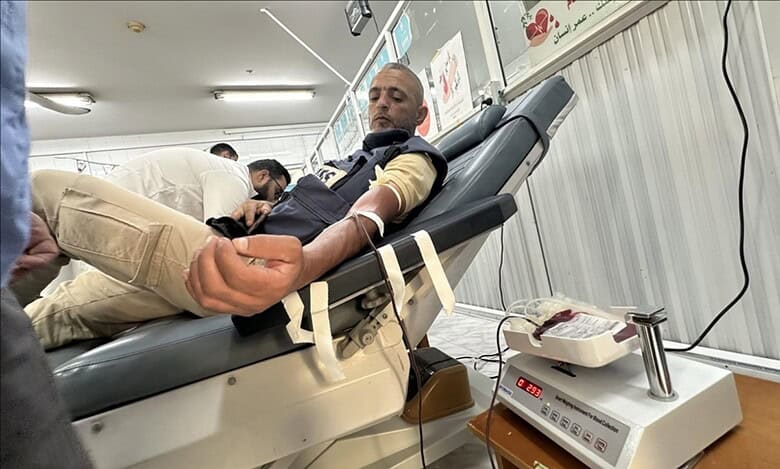 A Palestinian man with a beard lies reclined on a medical bed in a Gaza blood bank, donating blood through an IV in his arm; a nurse in white adjusts the tube connected to a blood collection bag on a scale, with Arabic health posters and heart symbols on the walls.