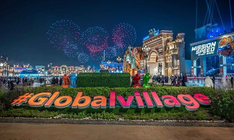A night view of Dubai’s Global Village with fireworks lighting up the sky, crowds walking around brightly lit pavilions, and a large #GlobalVillage sign in the foreground.
