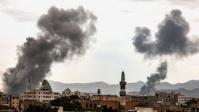 Smoke rises over buildings following an airstrike in Sanaa, Yemen, with dark plumes visible against a cloudy sky.