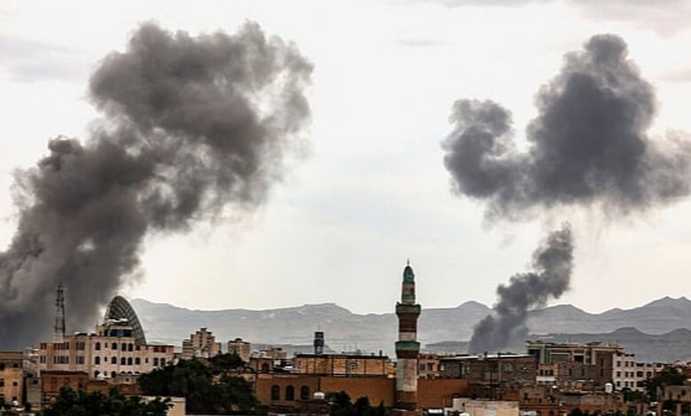 Smoke rises over buildings following an airstrike in Sanaa, Yemen, with dark plumes visible against a cloudy sky.