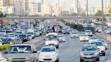 Busy multi-lane highway in Kuwait with many cars and SUVs during daylight, with city buildings in the background.
