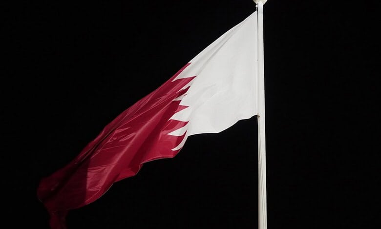 Qatar’s national flag waving against a dark night sky. The maroon-and-white flag is mounted on a tall pole.