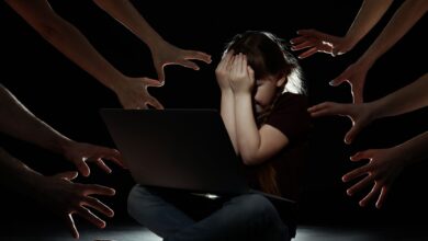 A young girl sits in front of a laptop, covering her face in distress, while numerous hands reach toward her from the darkness, symbolising online threats or cyberbullying.