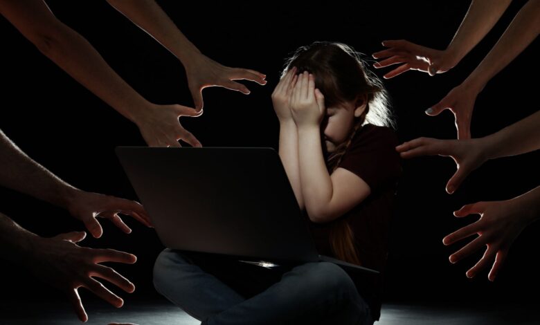 A young girl sits in front of a laptop, covering her face in distress, while numerous hands reach toward her from the darkness, symbolising online threats or cyberbullying.