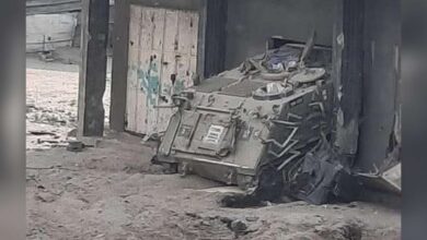 Booby-trapped armoured vehicle sits abandoned near a concrete building in Gaza City, surrounded by rubble and sand.