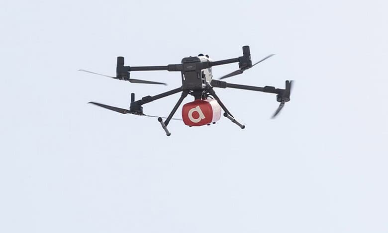 A delivery drone flying in the sky, carrying a red package with a white letter "a" on it.