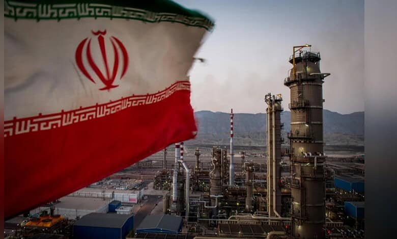 Iranian flag waving in the foreground with an oil refinery and industrial plant in the background.
