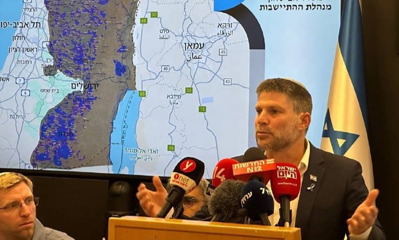 Bezalel Smotrich speaking at a press conference with a map of the West Bank behind him.