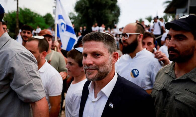 Israeli Finance Minister Bezalel Smotrich walking among a crowd with an Israeli flag in the background.
