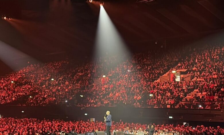 Mehdi Hasan speaking on stage at Wembley Arena during a Gaza fundraiser event, with a large audience in the background.