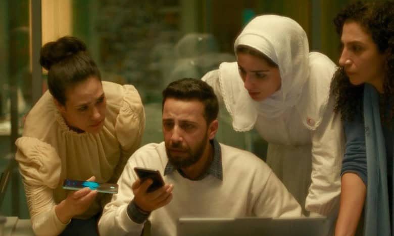 A tense scene showing three women and a man gathered closely, looking concerned at a phone screen