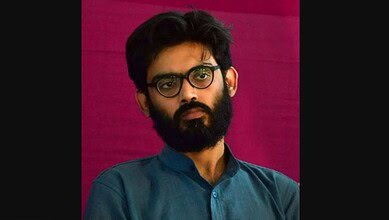 Young man with glasses and beard, wearing a blue shirt, posing against a pink background.