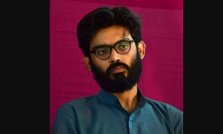 Young man with glasses and beard, wearing a blue shirt, posing against a pink background.