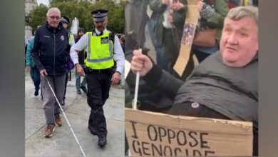 Two blind men at a London protest — one led away by police, another holding a sign reading “I oppose genocide.”