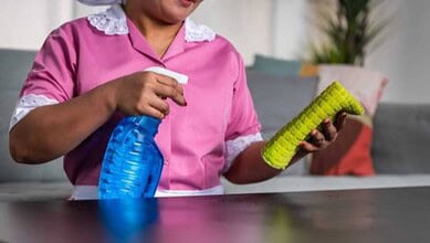 A domestic worker in uniform cleaning a table with a spray bottle and cloth.