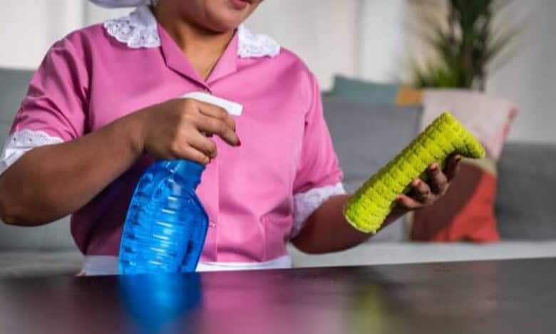 A domestic worker in uniform cleaning a table with a spray bottle and cloth.