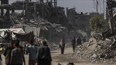 People walk through a street lined with destroyed buildings in a war-torn area.