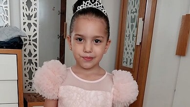 A young Palestinian girl, Hind Rajab, wearing a pink dress and tiara, smiles softly while standing indoors.