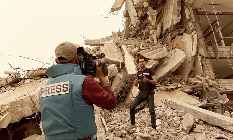 Two journalists wearing press vests report amid the rubble of a destroyed building in Gaza.