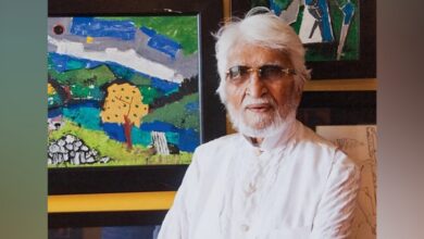 Indian painter MF Husain, dressed in a white kurta with a beard and white hair, wearing sunglasses, standing in front of colourful modern artworks.