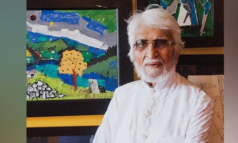 Indian painter MF Husain, dressed in a white kurta with a beard and white hair, wearing sunglasses, standing in front of colourful modern artworks.