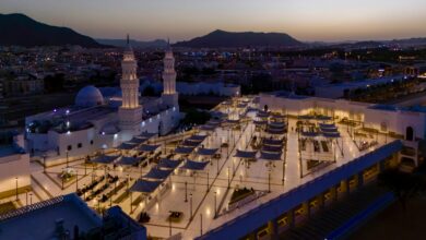 Aerial view of the illuminated Qiblatain Mosque in Madinah at dusk, showing its twin minarets and courtyard filled with worshippers.