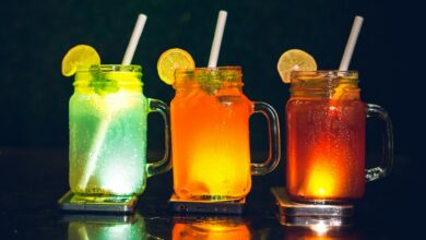 Three colourful sugar-sweetened drinks in glass jars with lemon slices and straws placed on a dark surface.
