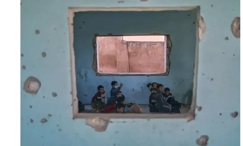 Children sit on the floor of a bullet-riddled classroom in Idlib, Syria, seen through a damaged wall.
