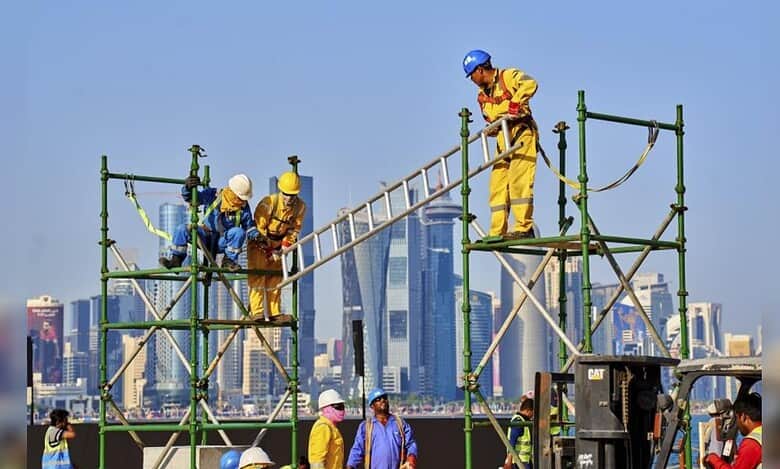 Migrant construction workers wearing safety gear and working on scaffolding with Gulf city skyscrapers in the background.
