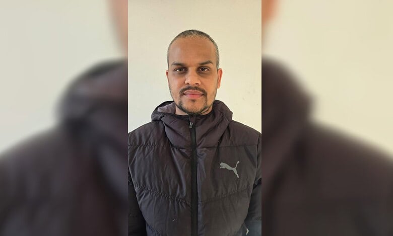 A man in a black winter jacket standing against a light-coloured wall.