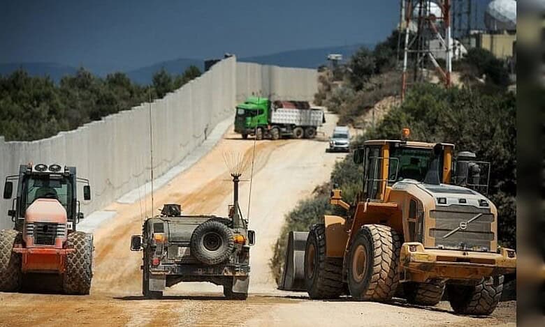Construction vehicles and a military jeep operate along a concrete border wall near a wooded area.