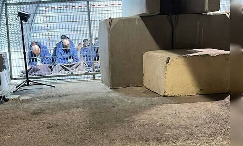 A group of blindfolded Palestinian detainees seated on the ground inside a fenced enclosure at the Sde Teiman military base, photographed under artificial lighting.