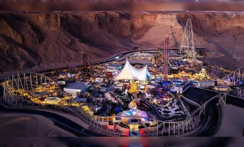 A night-time aerial view of Six Flags Qiddiya City, illuminated against the Tuwaiq Mountains with roller coasters and themed attractions.
