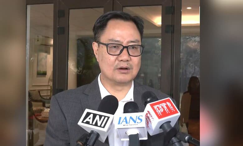 Kiren Rijiju in a dark suit and glasses speaking to media, with multiple microphones from ANI, IANS, and PTI news agencies positioned in front of him during a press conference