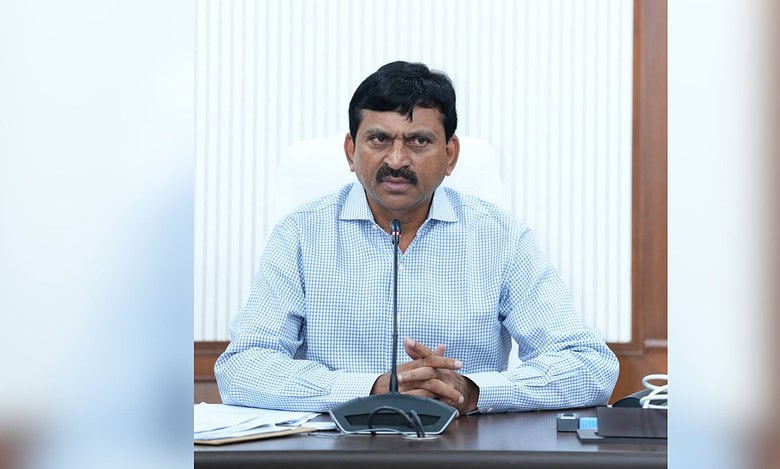 A man with a mustache wearing a light blue checkered shirt sits at a desk speaking into a microphone. He is gesturing with his hands while speaking. A conference phone and papers are visible on the desk in front of him, with a white curtained window in the background.