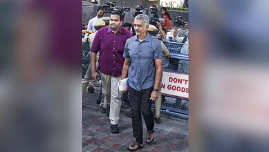 Two men walking on a street surrounded by media personnel with cameras, with police barricades visible in the background