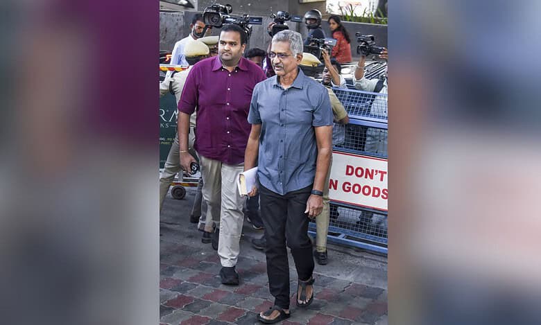 Two men walking on a street surrounded by media personnel with cameras, with police barricades visible in the background