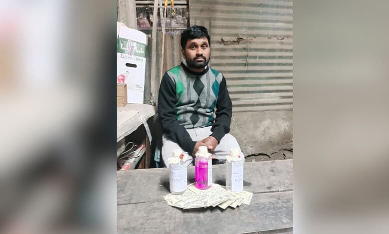 A man wearing a dark sweater with green and gray argyle pattern sits at a table with three bottles containing liquids (two clear, one pink) and Indian rupee notes arranged in front of him. The setting appears to be a small shop or workspace with corrugated metal walls and shelving visible in the background