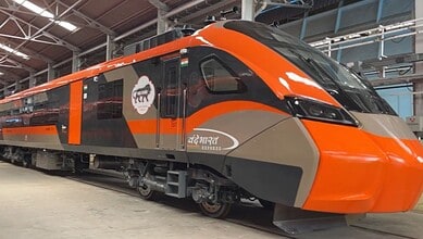Image shows Modern orange and gray Vande Bharat Express train in maintenance facility