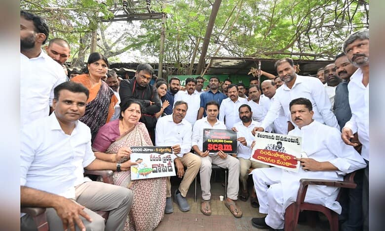 Political gathering showing BJP leaders and supporters sitting outdoors holding protest signs in Telugu about alleged scams. Group photo with men in white attire and a woman in saree, taken under trees with makeshift shelter in background.