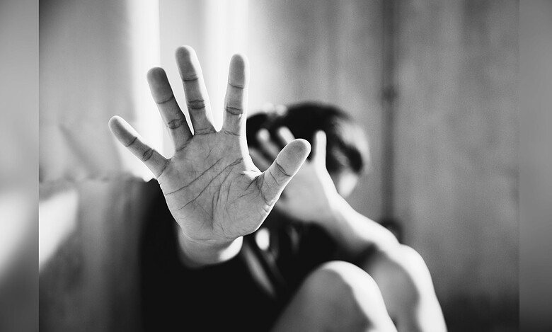 A person holds up their hand toward the camera in a defensive "stop" gesture while their face is obscured in the blurred background, conveying distress or a plea for help.