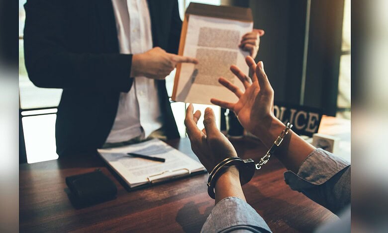A person in handcuffs sits at a table during what appears to be a legal consultation, while someone in professional attire stands nearby holding a document. Law books and papers are visible on the wooden table surface.
