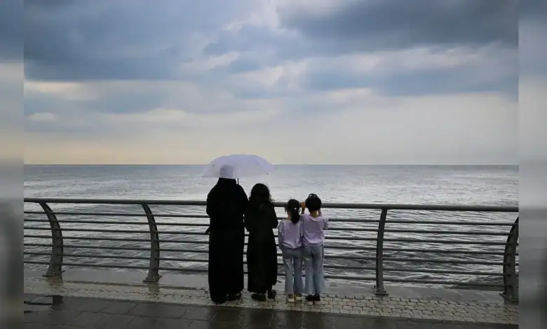 A woman and three children stand by the Jeddah waterfront under cloudy skies, looking out at the sea while holding an umbrella.