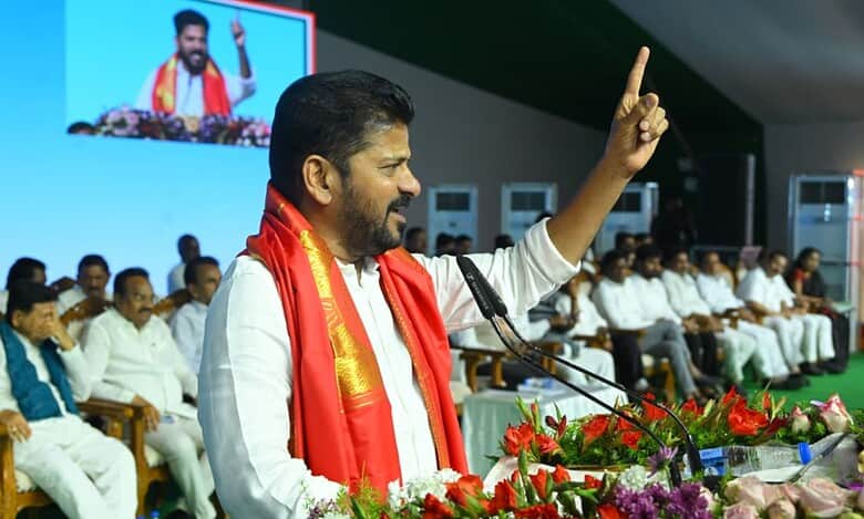 Political leader in white attire with red traditional shawl speaking at podium with raised finger gesture, addressing audience seated in background, with large screen display and floral arrangements visible