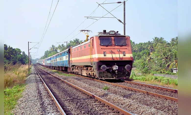 An orange and yellow electric locomotive pulling blue passenger coaches through lush green countryside on an electrified railway track