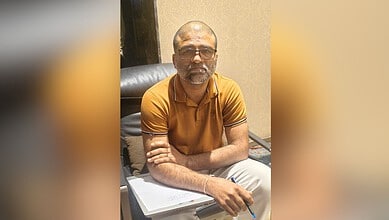 Image shows Middle-aged man wearing glasses and orange polo shirt sitting at desk with pen and notebook