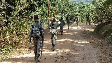 Image shows Armed soldiers in camouflage uniforms walking along a dirt road through a rural, tree-lined area.