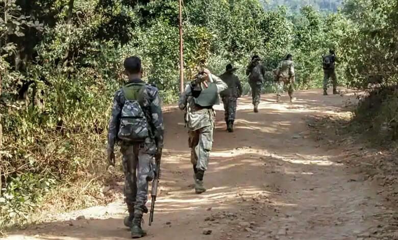 Image shows Armed soldiers in camouflage uniforms walking along a dirt road through a rural, tree-lined area.