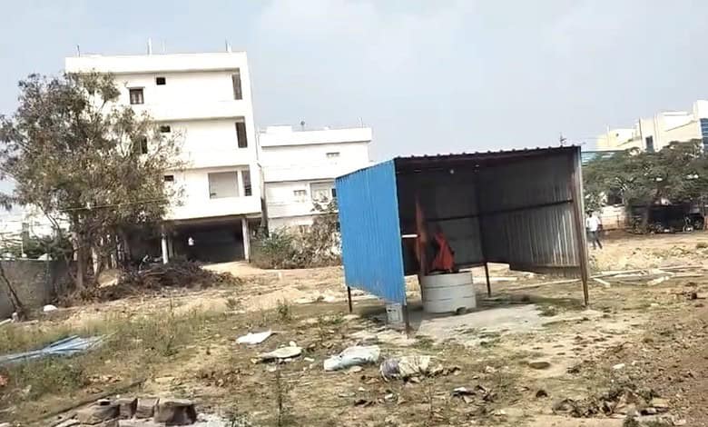 Image shows a vacant urban plot with makeshift shed and residential buildings in background