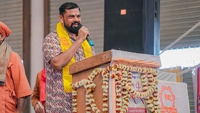 Raja Singh speaking at a rally with a microphone, wearing a yellow scarf, during a political gatheri.